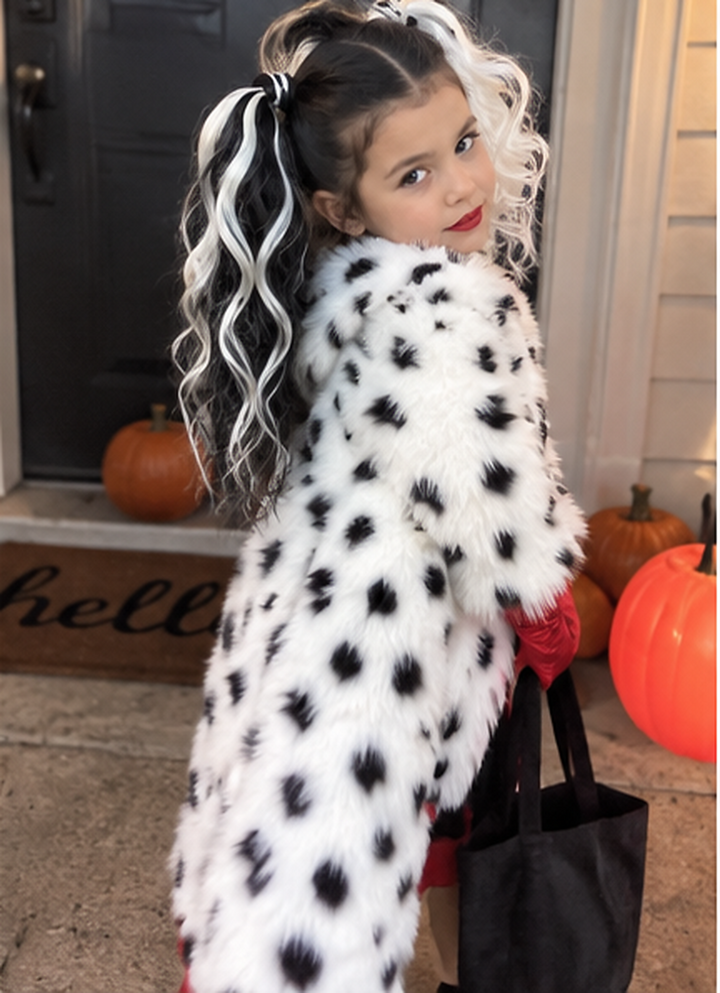 A young girl wearing a white faux fur coat with black spots, styled like a Dalmatian, standing on a front porch with pumpkins and holding a black tote bag.