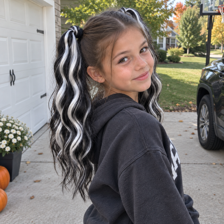 A young girl with long brown hair styled in two high pigtails, one black with white streaks and the other black with light brown streaks, wearing a dark gray hoodie. She is standing in a driveway in front of a white garage door and a green lawn.
