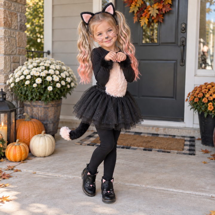 A young girl wearing a black and pink cat costume stands on a front porch. The costume includes a black long-sleeve top, black tulle tutu skirt, black leggings, a fluffy light pink belly patch, and a black tail with a pink pom-pom.
